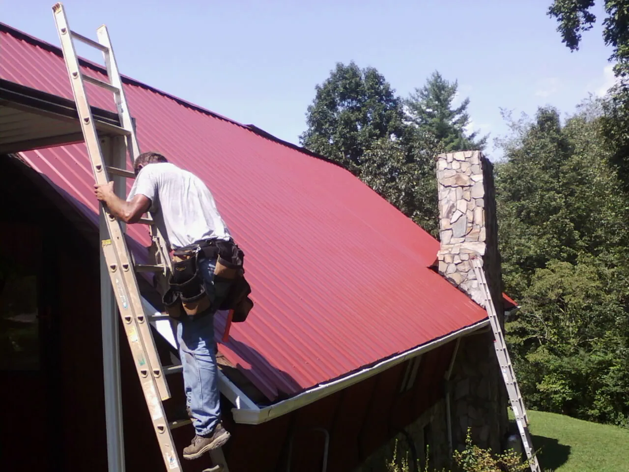 Roofer on a ladder beside a red metal roof and stone chimney — Soffit & Fascia Repair by Roofing Brothers in Grand View